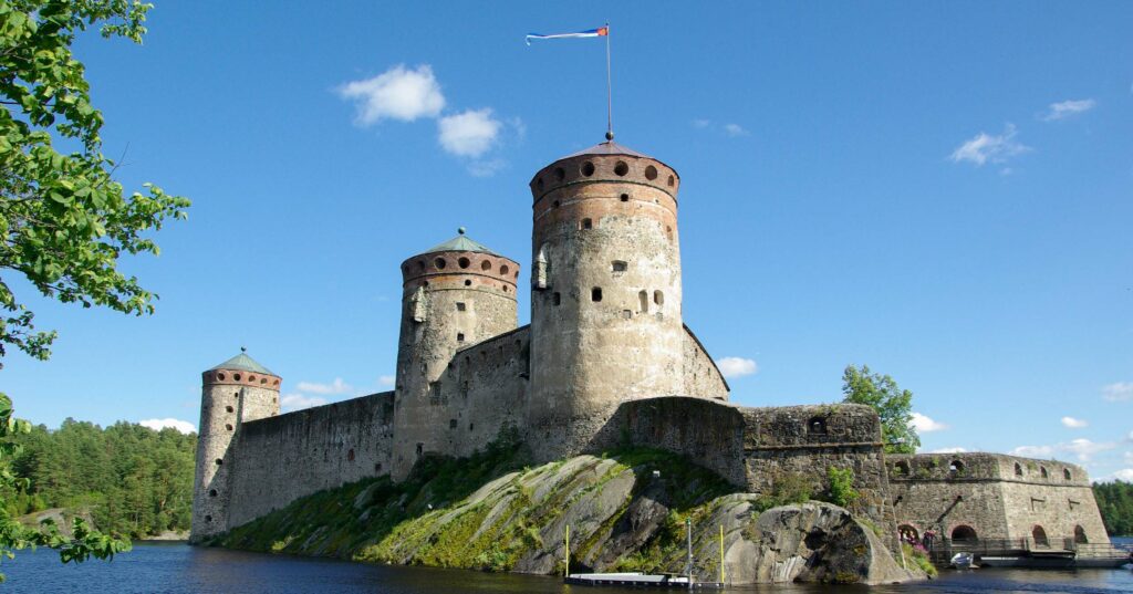 A majestic ancient stone castle with cylindrical towers, situated on a rocky landscape surrounded by water, under a clear blue sky with lush green trees in the foreground