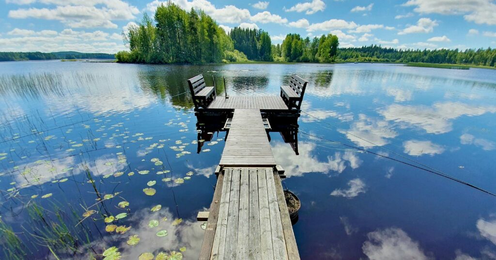 A serene view of a wooden dock with two chairs, extending over a calm lake surrounded by greenery under a partly cloudy sky