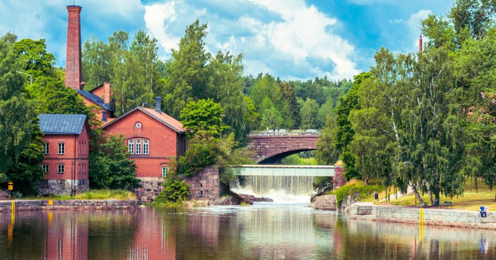 A serene landscape featuring a red brick building with a tall chimney, situated beside a calm lake with a waterfall and an arched stone bridge in the background, surrounded by lush green trees under a partly cloudy sky