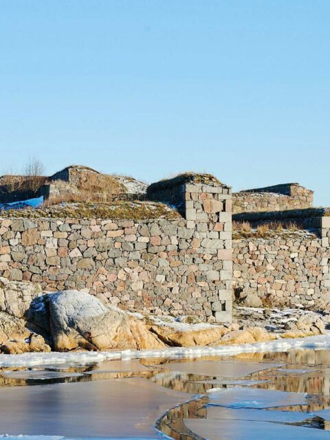 winter landscape featuring a stone structure on the snowy and rocky shore, with a partially frozen body of water in the foreground and clear blue skies above