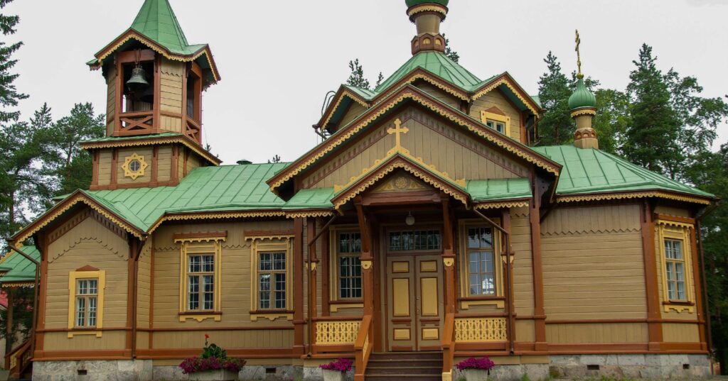 A wooden orthodox church with green roofs and a bell tower, surrounded by trees