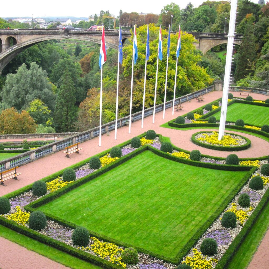 A colorful garden with blooming flowers and plants. Flags of various countries flutter in the background, and a bridge can be seen in the far distance