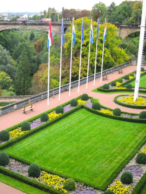 A colorful garden with blooming flowers and plants. Flags of various countries flutter in the background, and a bridge can be seen in the far distance