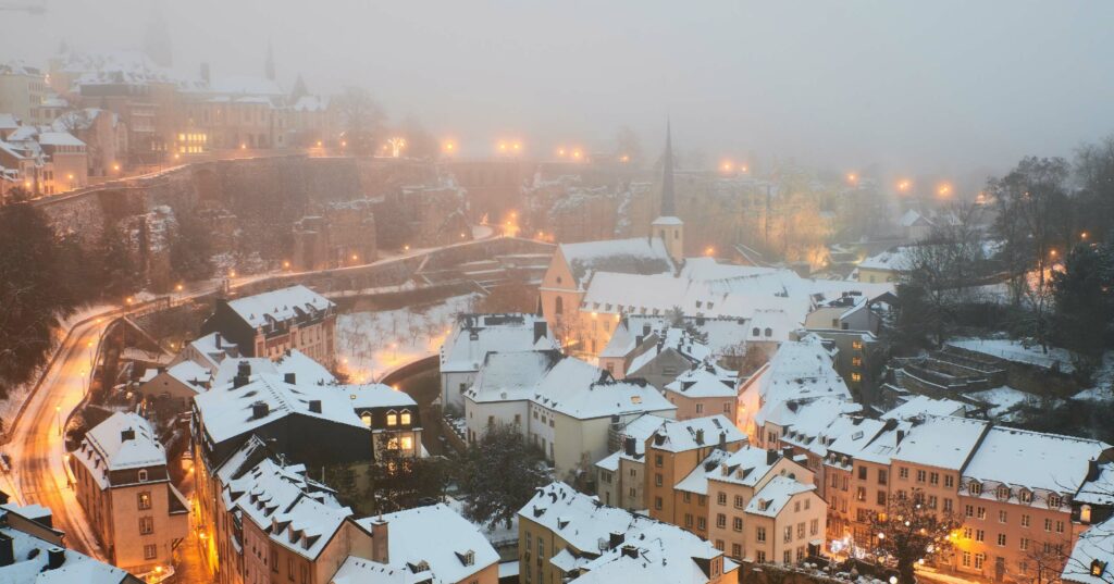 snow-covered city with warm lights and dark trees in foreground