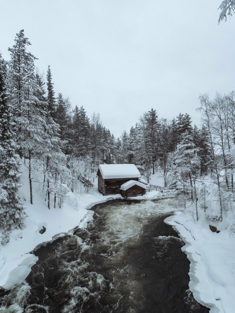 A serene winter landscape featuring a snow-covered cabin beside a flowing river, surrounded by frosty trees