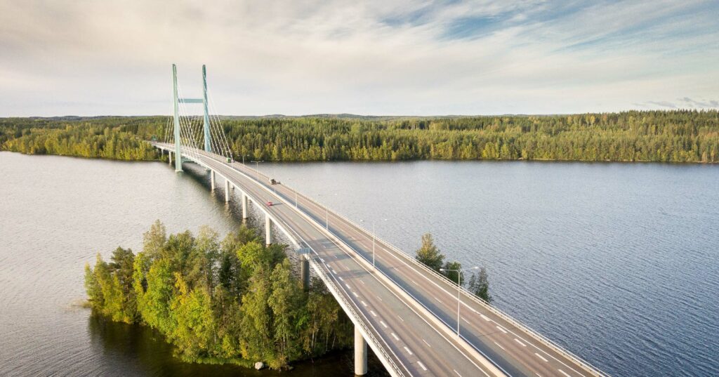 A long bridge with white supports crossing a calm lake, surrounded by lush greenery under a clear sky