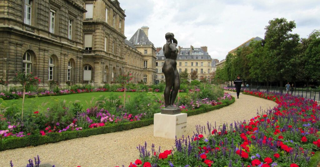 A statue of a figure stands prominently in a beautifully landscaped garden, with an elegant stone building in the background